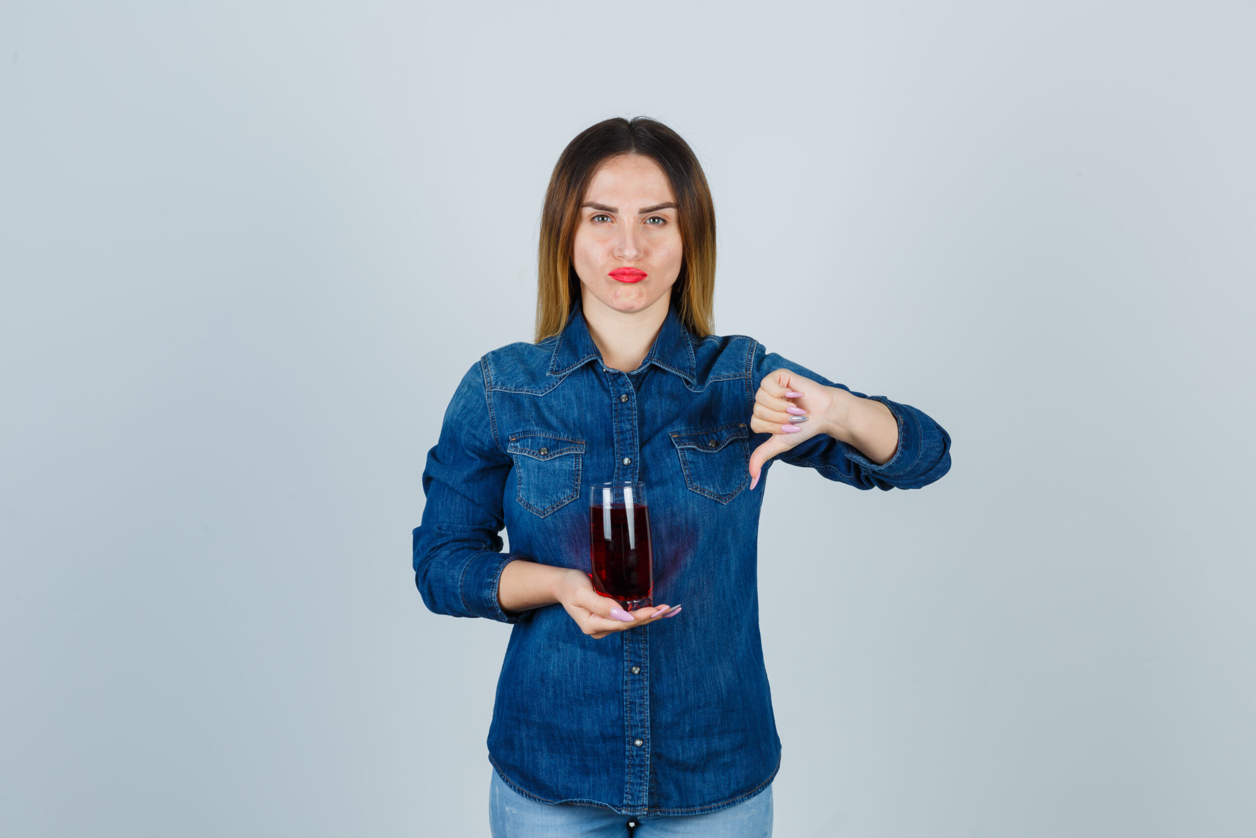 young girl holding glass of red liquid in one hand, showing thumb down in jean shirt, pants and looking displeased , front view.